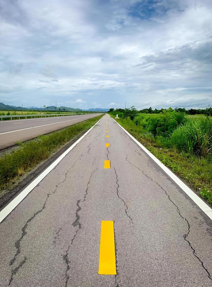 Perspective cracked asphalt road ahead to the mountain. Long straight damaged asphalt road. Bad surface of bike lane and white sky and clouds with green grass beside the empty road. Nature landscape. photo