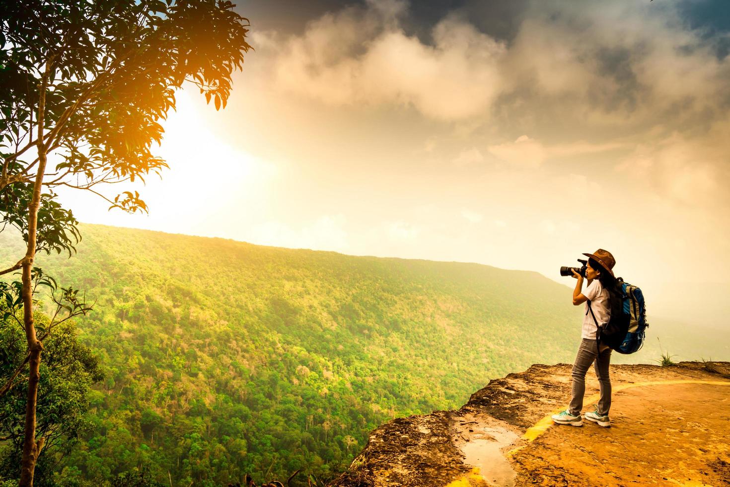 Young traveling woman with backpack, hat and camera stand on the mountain cliff. Female photographer taking picture of tropical forest, sky and clouds on her vacation. Asian woman travel alone. photo