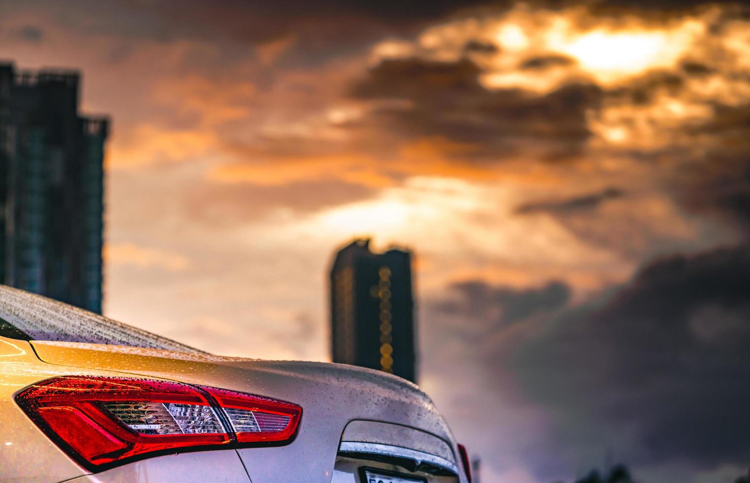 Rear view of car parked in the city near high building after rain with grey and orange sky and clouds and have water droplet on surface of car. Hybrid auto concept. Travel in the city on rainy season. photo
