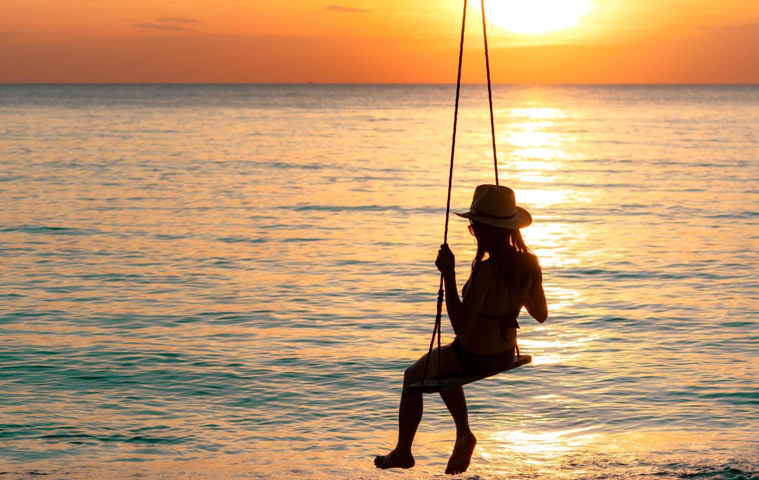 Silhouette woman wear bikini and straw hat swing the swings at the beach on summer vacation at sunset. Enjoying and relaxing girl on holiday. Summer vibes. Woman watching beautiful sunset sky. photo