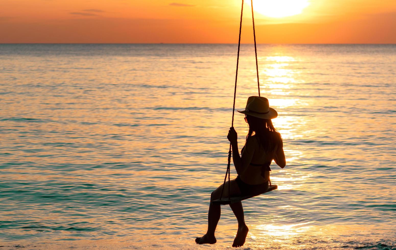 silueta mujer usa bikini y sombrero de paja columpios en la playa en vacaciones de verano al atardecer. disfrutando y relajando a una chica de vacaciones. vibras de verano. mujer mirando el hermoso cielo del atardecer. foto