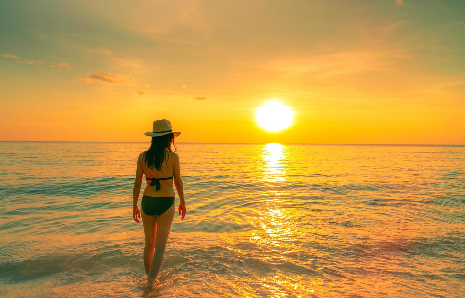 silueta mujer sexy caminando en el mar tropical con hermoso cielo al atardecer en la playa del paraíso. niña feliz usar bikini y sombrero de paja relajantes vacaciones de verano. viajes de vacaciones vibras de verano. la vida continua. foto