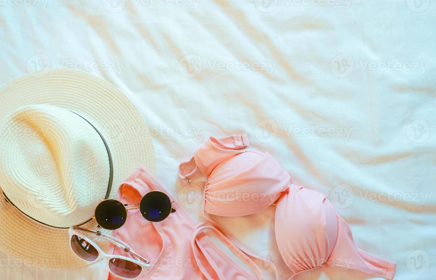 Top view of bikini swimsuit, sunglasses, and straw hat on bed sheet. Woman's swimwear and beach accessories on bed of resort. Pink bikini swimwear set for the beach. Summer vacation and summer vibes. photo