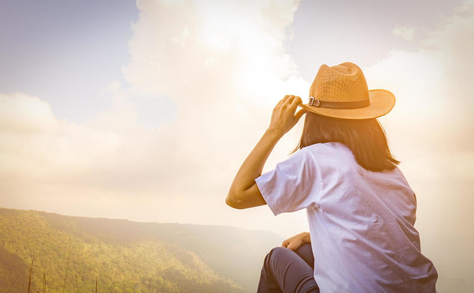 Young traveling woman wearing hat and sitting on the top of the mountain cliff with relaxing mood and watching beautiful view of woods and blue sky and clouds on vacation. Asian woman travel alone. photo