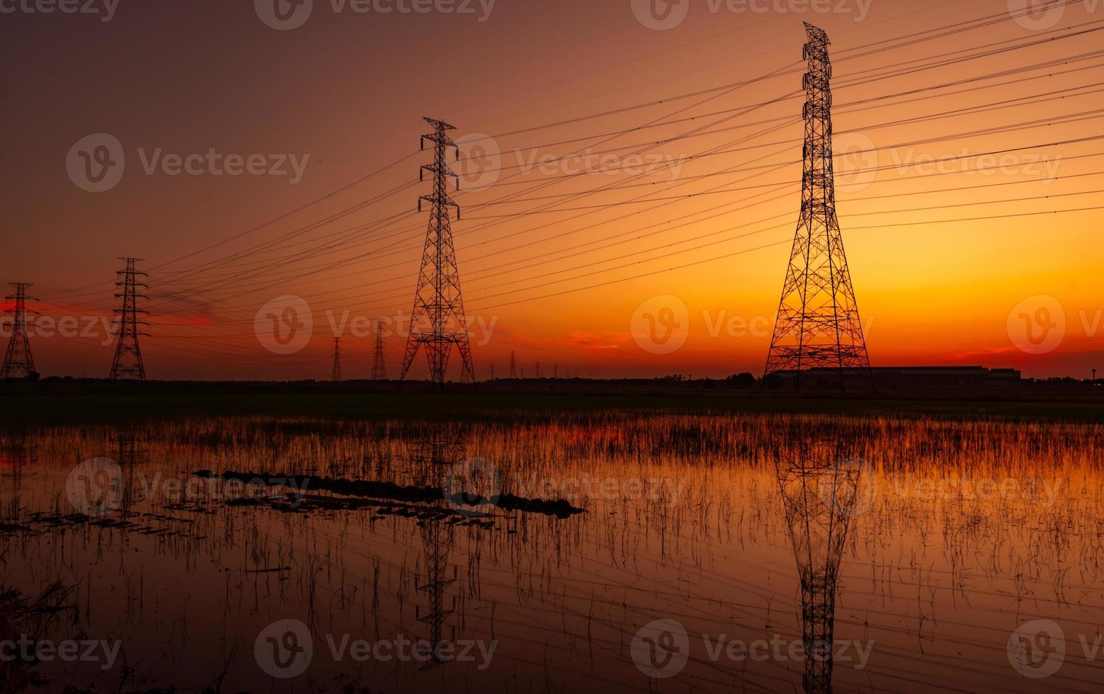 High voltage electric pylon and electrical wire with sunset sky. Electricity poles. Power and energy concept. High voltage grid tower with wire cable at rice farm field near industrial factory. photo