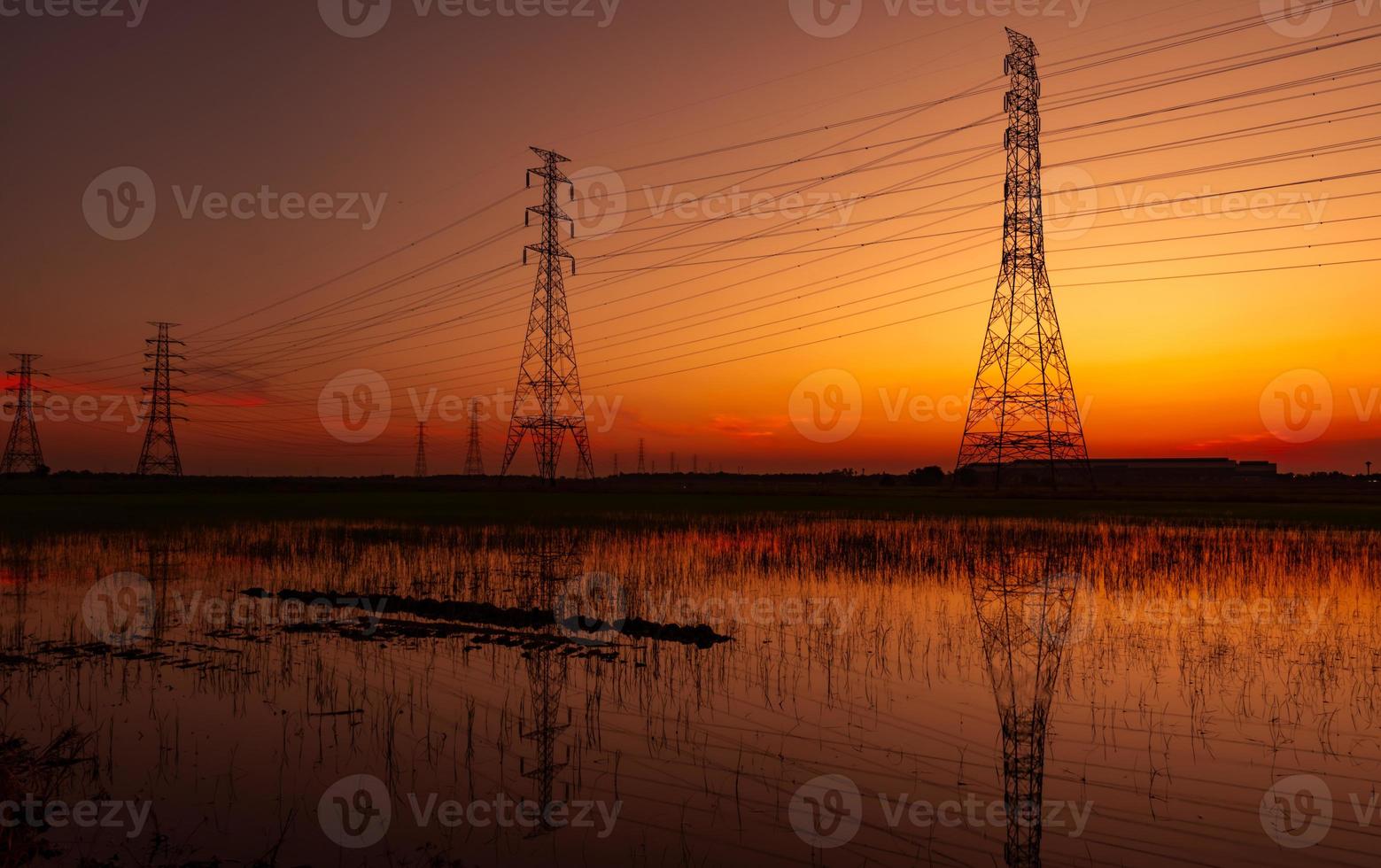 pilón eléctrico de alto voltaje y cable eléctrico con cielo al atardecer. postes de electricidad concepto de potencia y energía. torre de red de alto voltaje con cable de alambre en el campo de cultivo de arroz cerca de la fábrica industrial. foto