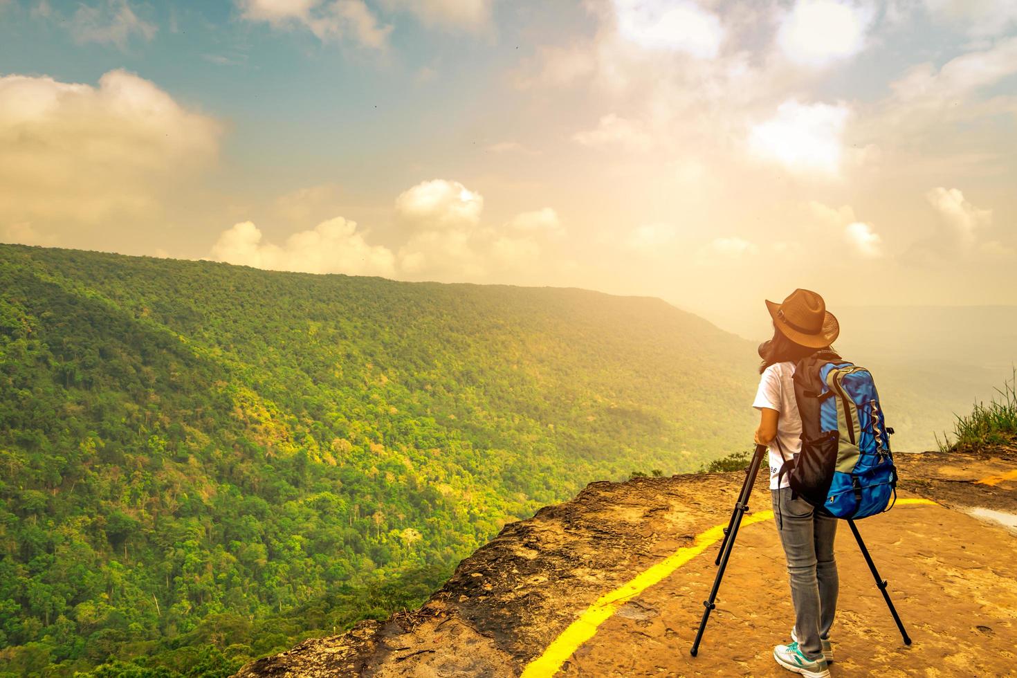 Young traveling woman with backpack hat and camera on tripod stand on the top of the mountain cliff watching beautiful view of woods and sky on her vacation. Asian woman travel alone. photo