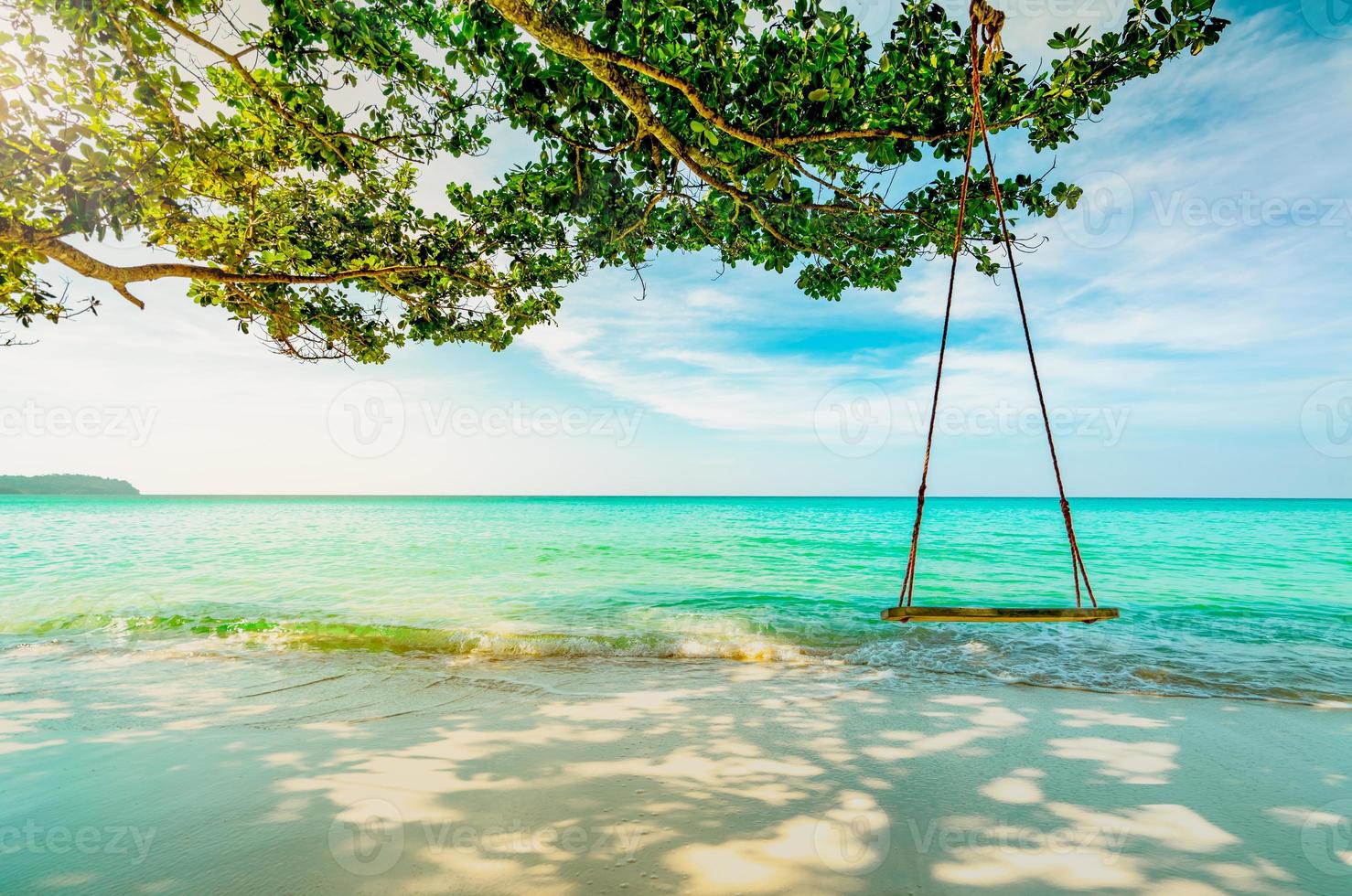 Wooden swings hang from branch of tree at seaside. Emerald green sea water with blue sky and white clouds on summer. Summer vibes. Summer vacation. Empty swings at sand beach with morning sun light . photo