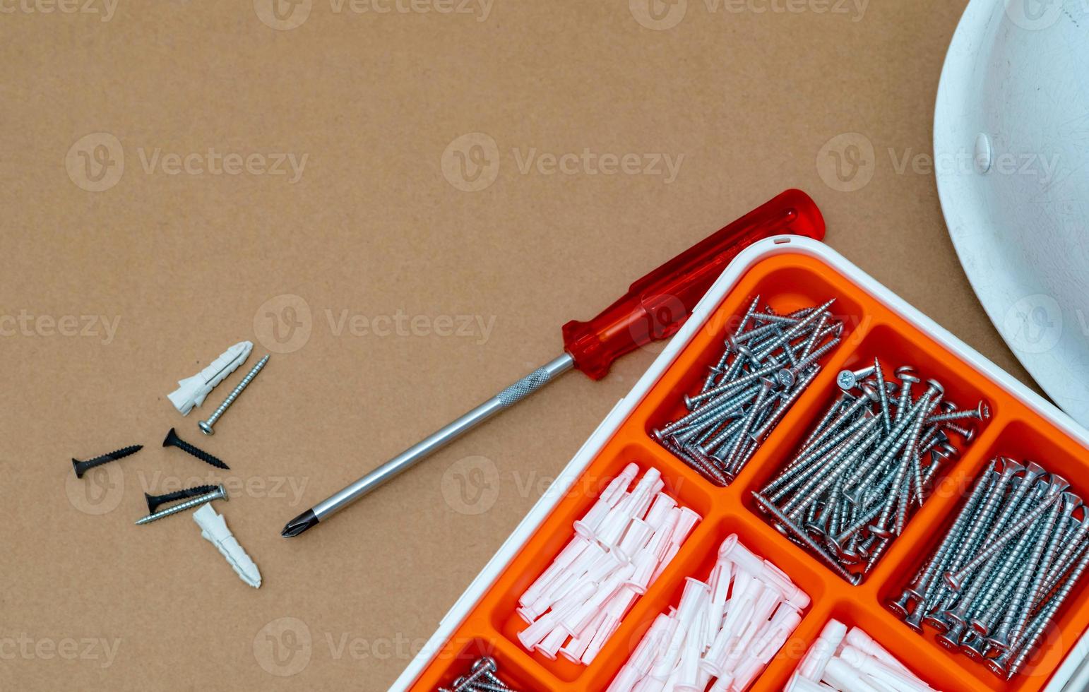 Top view of plastic plug and screw in white and orange plastic box. Screwdriver, gray wall plug, black screw, and safety helmet on brown paper box texture. Tools set for installation and construction. photo