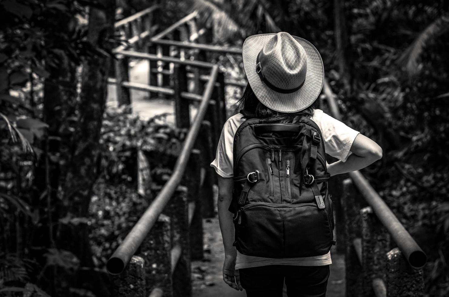 Black and white photo of Asian woman tourist with hat and backpack standing and start walking on nature trail bridge in evergreen forest. Alone young woman traveler.