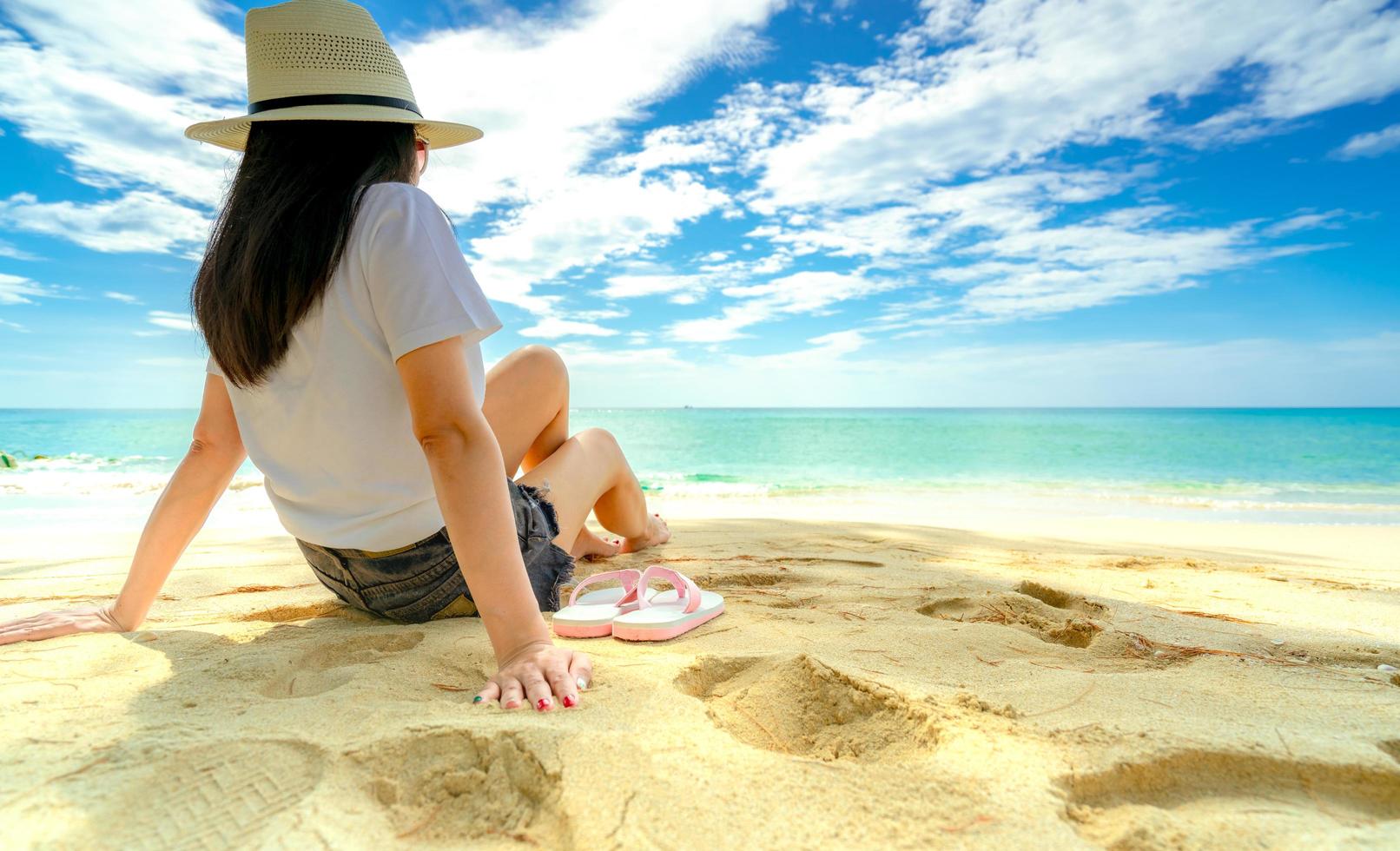 mujer joven feliz en camisas blancas y pantalones cortos sentados en la playa de arena. relajarse y disfrutar de unas vacaciones en la playa del paraíso tropical con cielo azul y nubes. chica en vacaciones de verano. vibras de verano. día feliz. foto