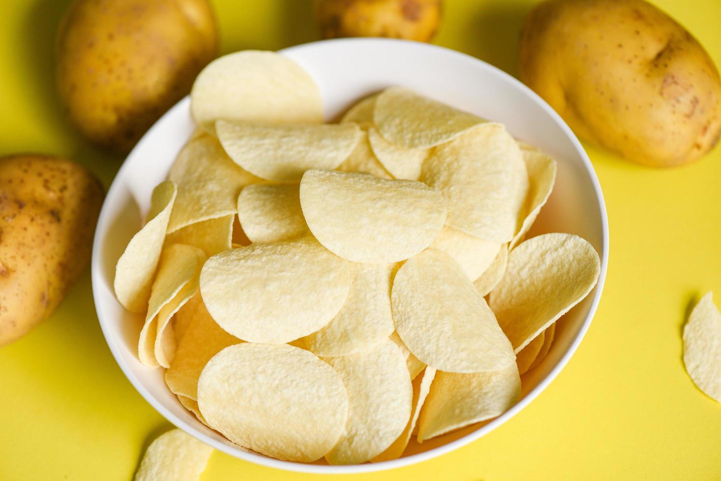 Crispy potato chips on the kitchen table and fresh raw potatoes on yellow background, Potato chips snack on bowl photo