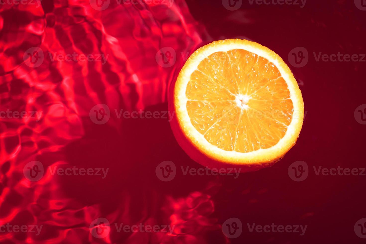 Piece of fresh orange on water surface with waves and reflections. Swimming piece of orange on red abstract background. photo