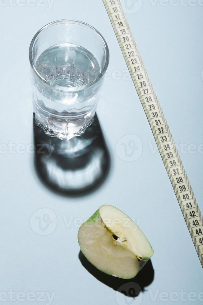 Healthy livestyle diet concept layout. Glass of water with apple slice and a tape measure on blue background. photo