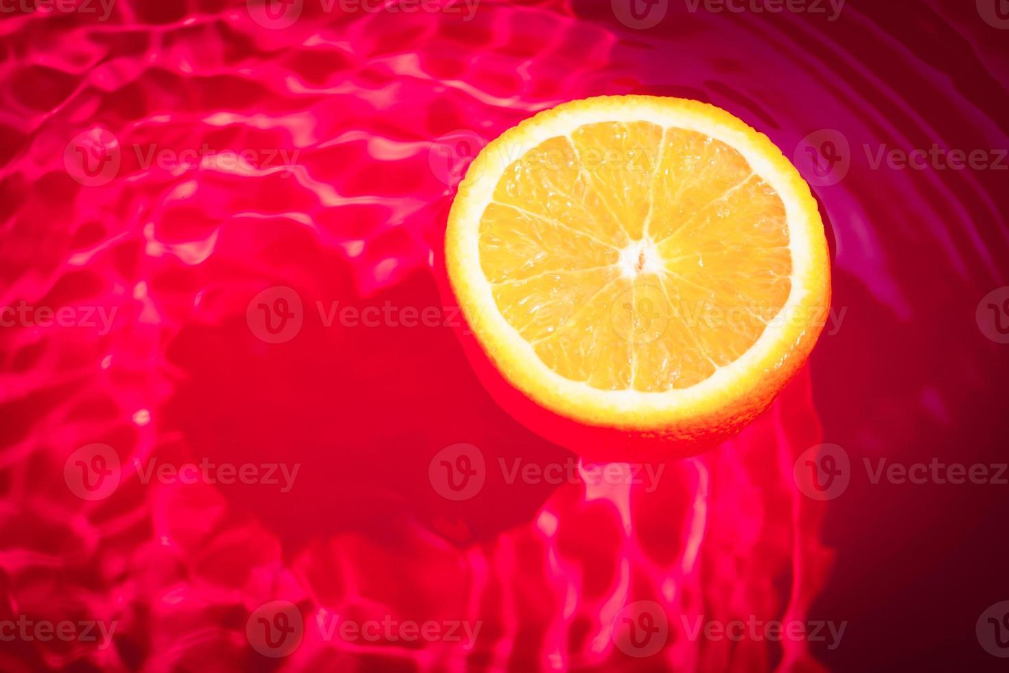 Piece of fresh orange on water surface with waves and reflections. Swimming piece of orange on red abstract background. photo