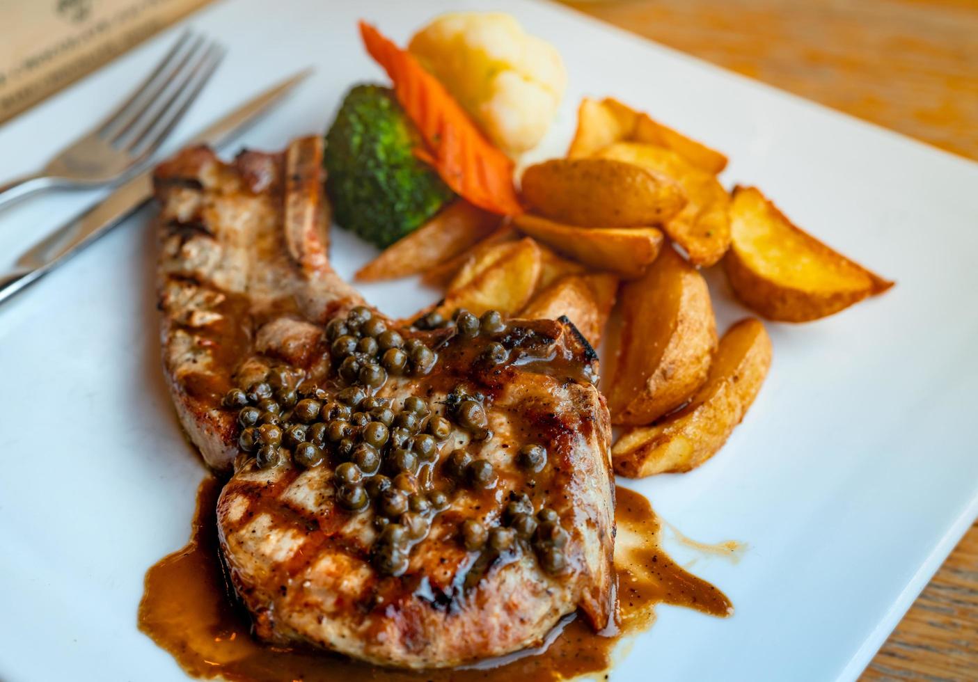 Steak topped with sauce and pepper serve with potato fried and carrots, cauliflower and broccoli steamed. Steak on white plate with stainless steel fork and knife put on wooden table. Tomahawk steak. photo