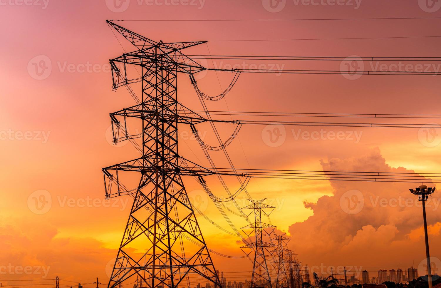 torres eléctricas de alto voltaje y líneas de transmisión con cielo de puesta de sol naranja y fondo de edificio de oficinas moderno de paisaje urbano. torres de electricidad. potencia y energía para la ciudad. torre de red de alta tensión. foto