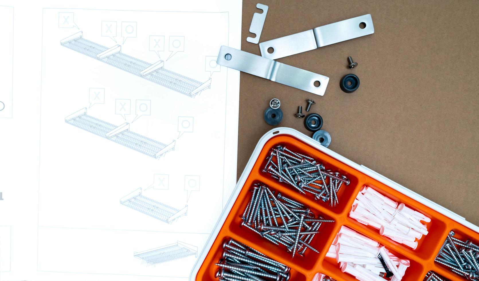 Screws and wall plug in orange plastic box. Top view tool box on brown background with dish drying rack assembly instruction. Set of screws and plastic plug for drilling wall and installation things. photo