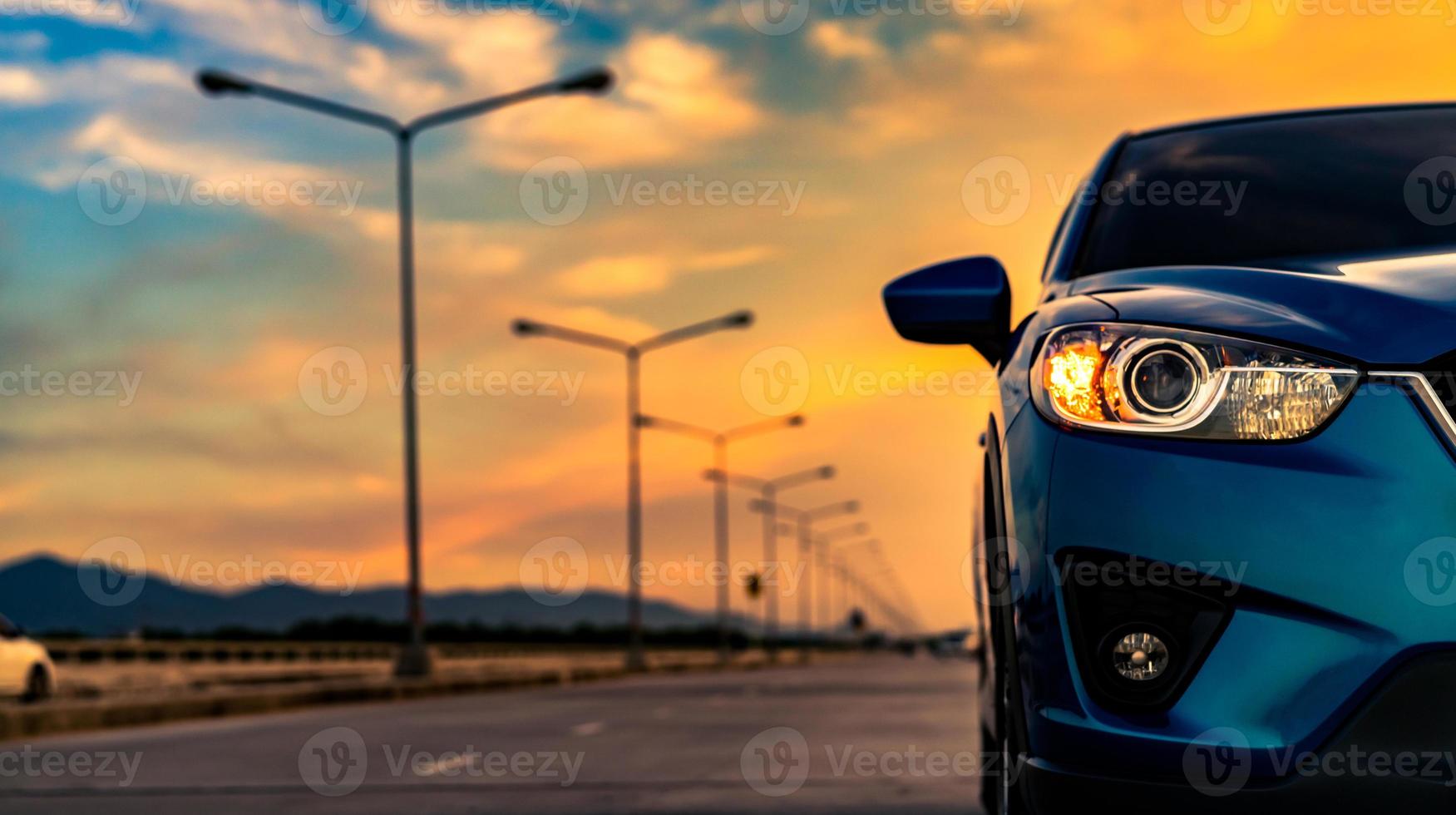 Blue compact SUV car open headlamp light parked on concrete road near the mountain at sunset with beautiful sky and clouds. Road trip travel. Automotive industry. Electric and hybrid car technology. photo