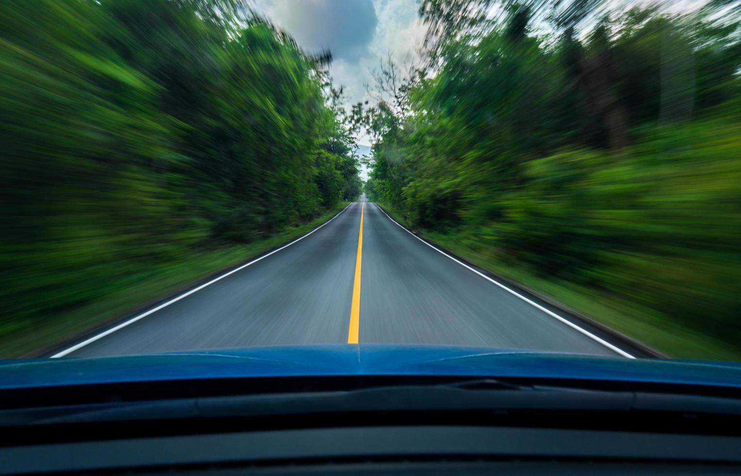 Front view of blue car driving with fast speed on the middle of asphalt road with white and yellow line of traffic symbol in the green forest. Trees beside the road is blurred. Car with blur motion. photo