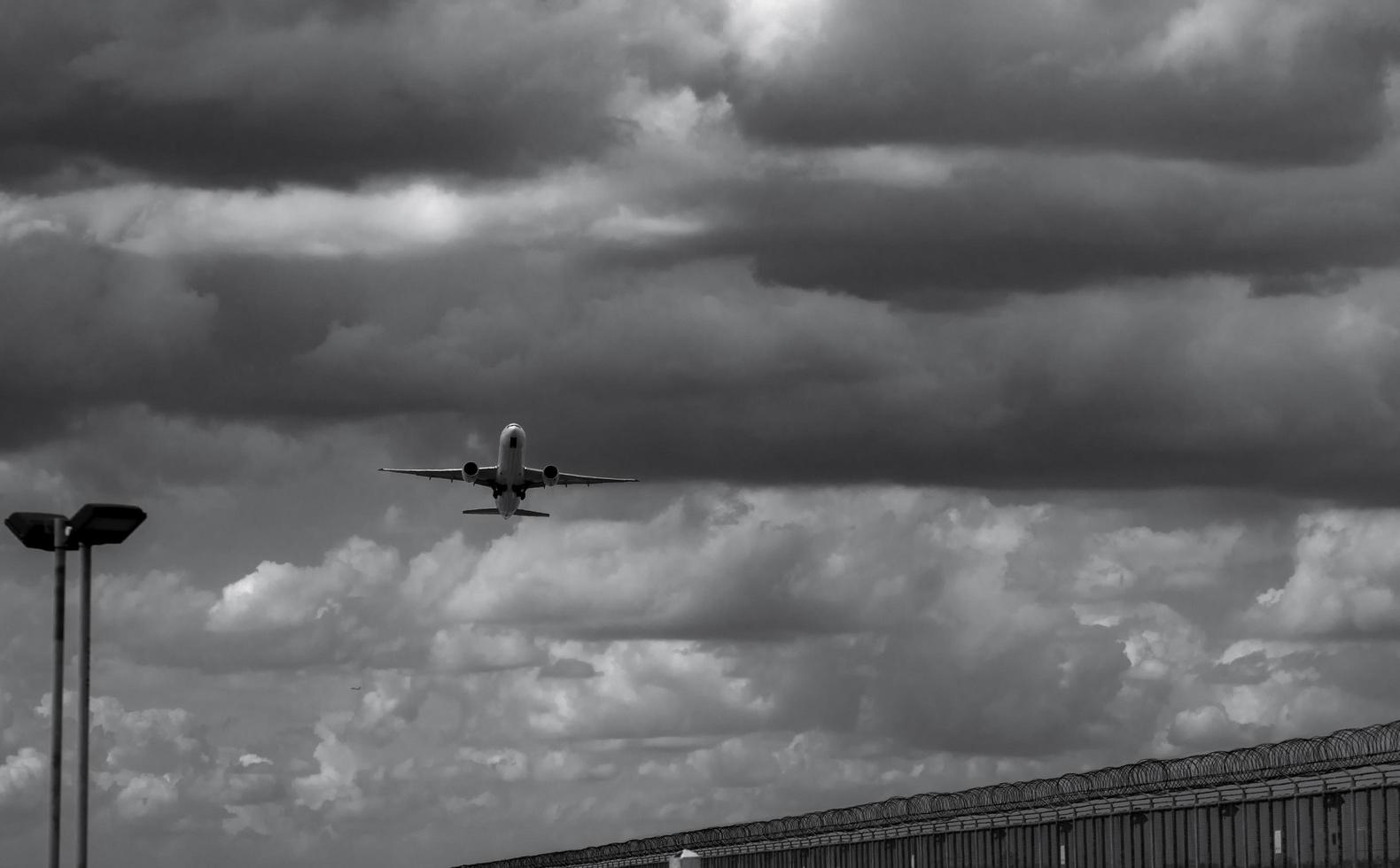 Black and white scene of airplane landing at the airport. Commercial airline flying on dark sky and white clouds over fence and lamp post. Aviation business crisis concept. Journey vacation flight. photo