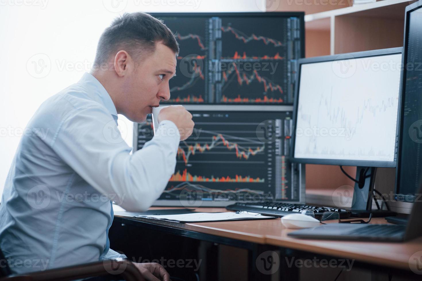 Drinks some coffee to stay concentrated. Man working online in the office with multiple computer screens in index charts photo