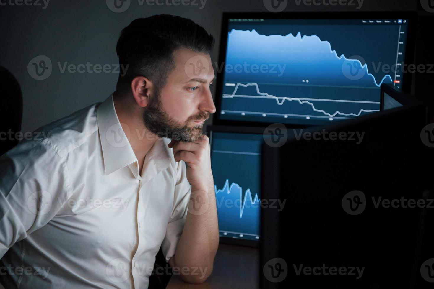 siguiendo los cambios en el mercado. hombre barbudo con camisa blanca trabaja en la oficina con múltiples pantallas de computadora en gráficos de índice foto