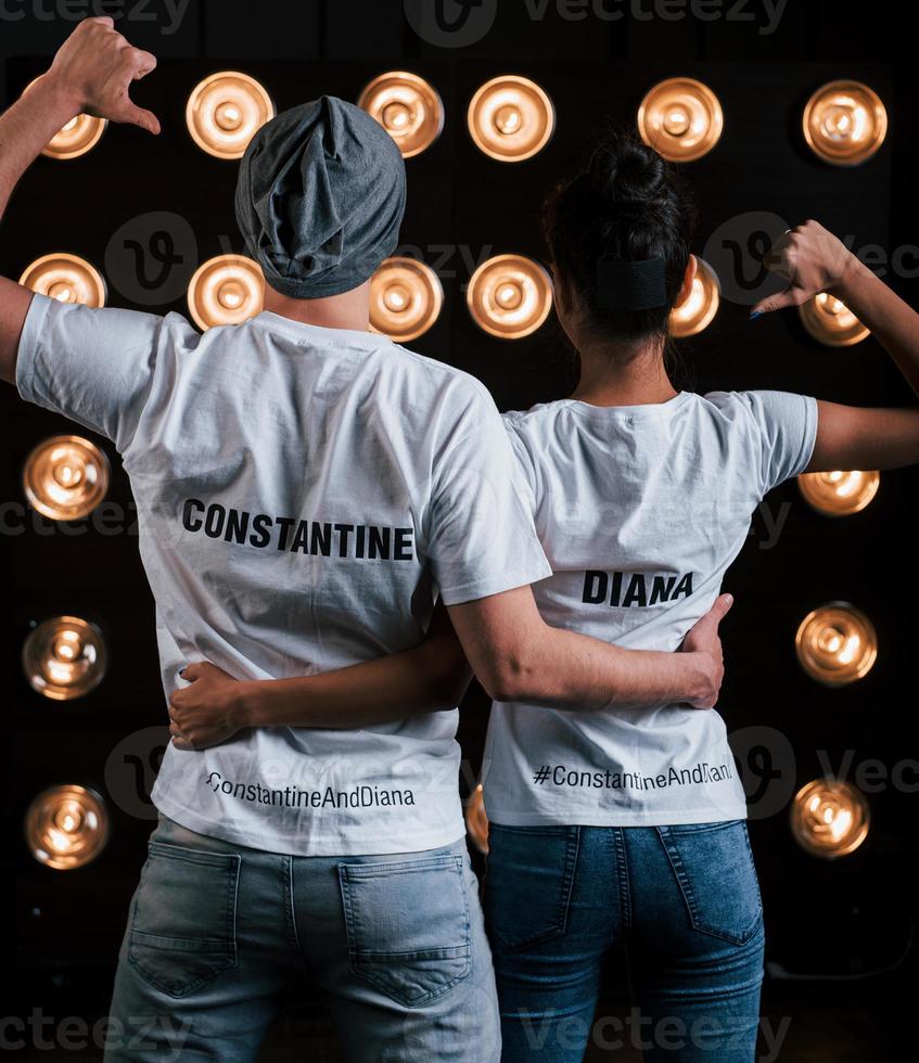 Shows thumbs on themselves. Back view of couple in shirts with their names on it standing next to studio lights photo