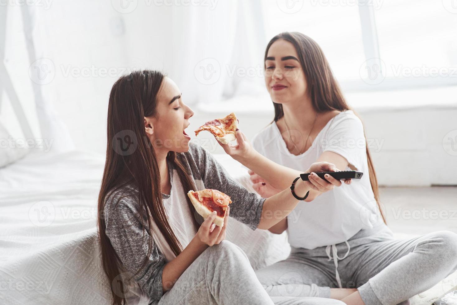 Taste mine slice. Sisters eating pizza when watching TV while sits on the floor of beautiful bedroom at daytime photo