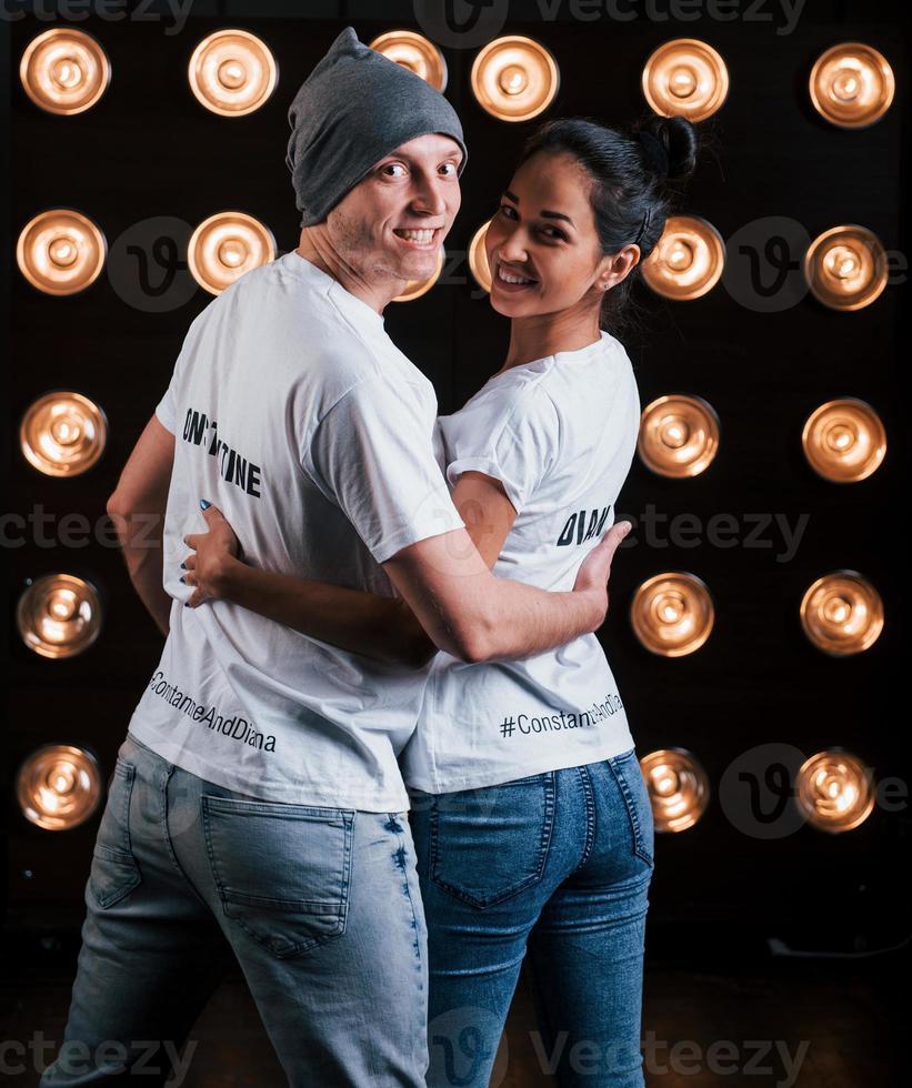 Turned to the camera. Back view of couple in shirts with their names on it standing next to studio lights photo