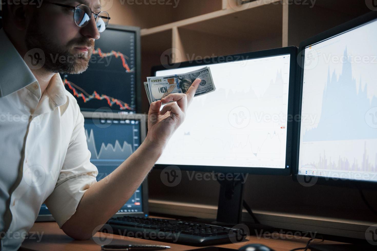 con efectivo en la mano. hombre barbudo con camisa blanca y anteojos tiene dinero en la oficina con múltiples pantallas de computadora en gráficos de índice foto