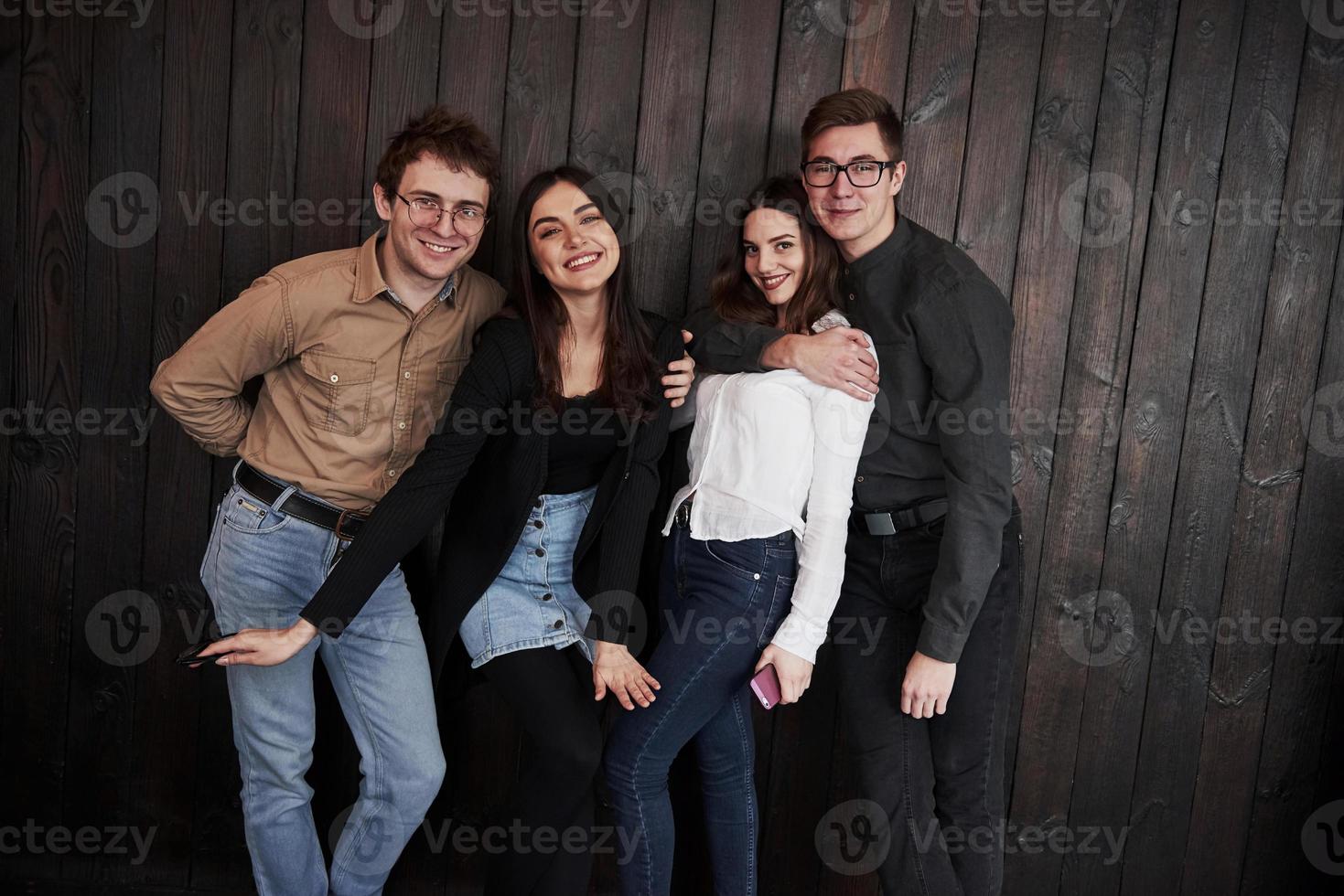 Guy hugs girl. Youth stands against black wooden wall. Group of friends spending time together photo