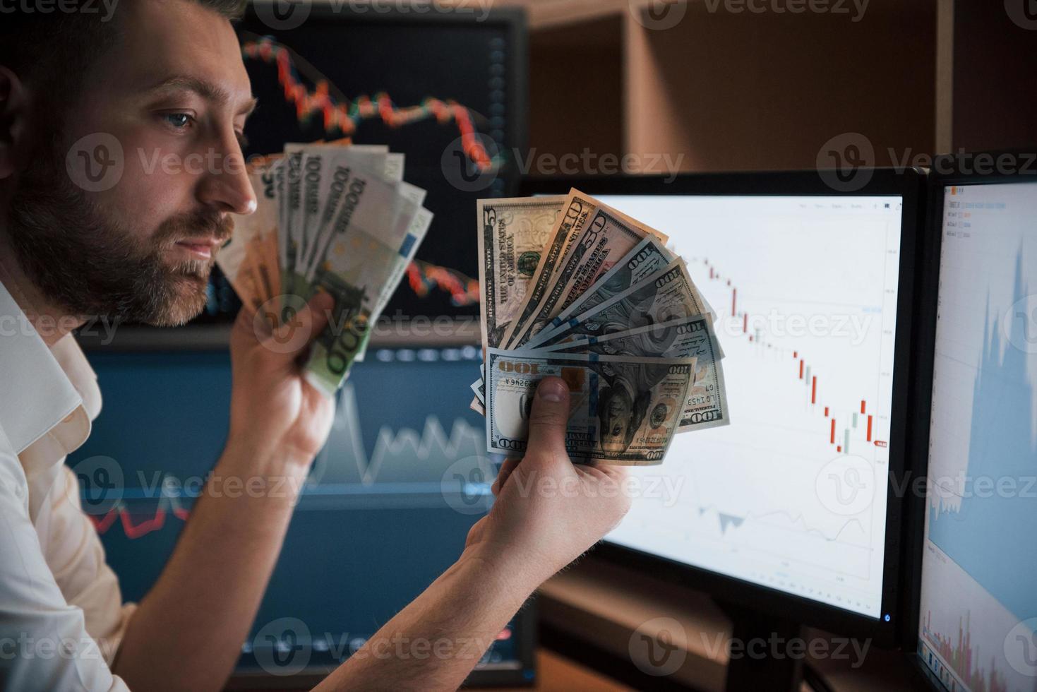 Rich person. Bearded man in white shirt holds earned money in the office with multiple computer screens in index charts photo