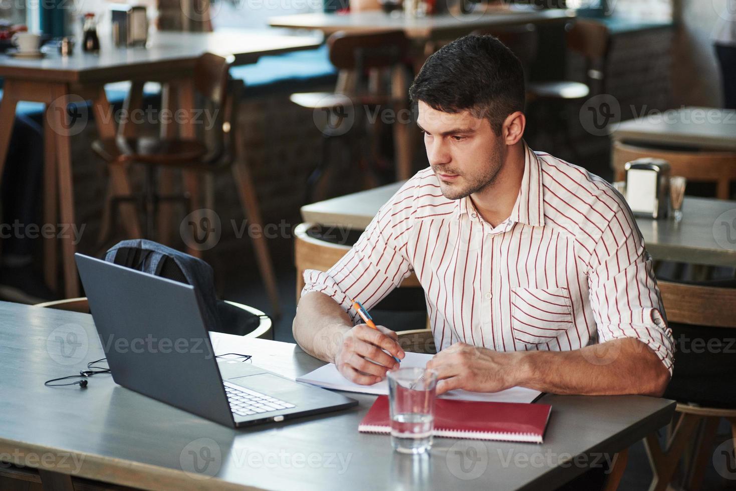 Busy guy looks at monitor and writes tasks for his business plan. Adult man sits in cafe at daytime and using the laptop for the remote work photo