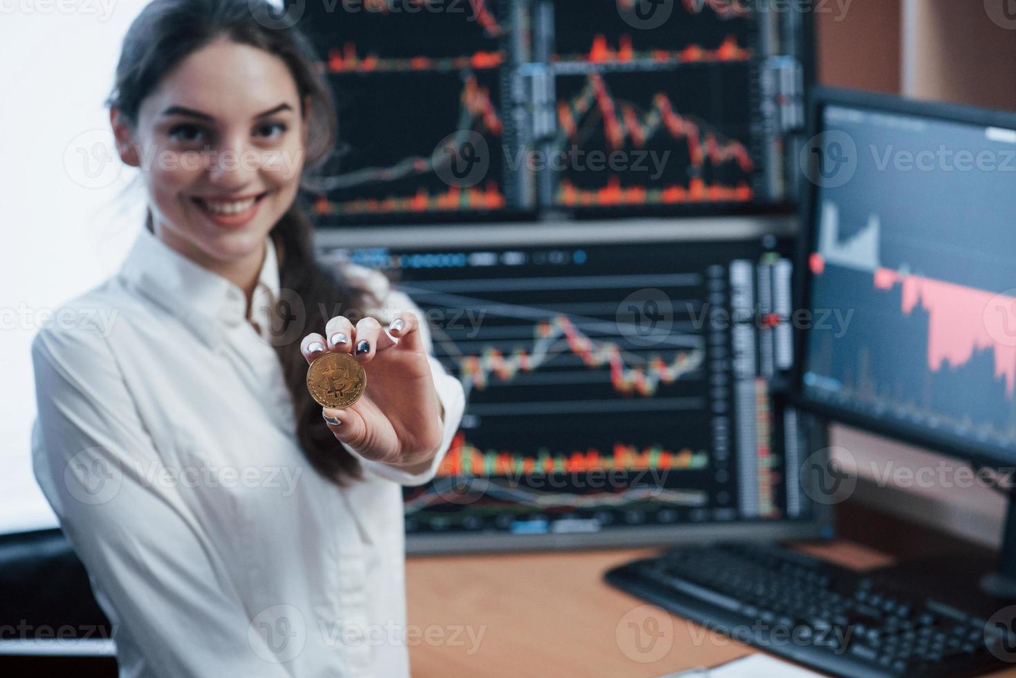 Nice daylight. Beautiful woman holding bitcoin in hands while standing in modern office with many monitors with graphs photo