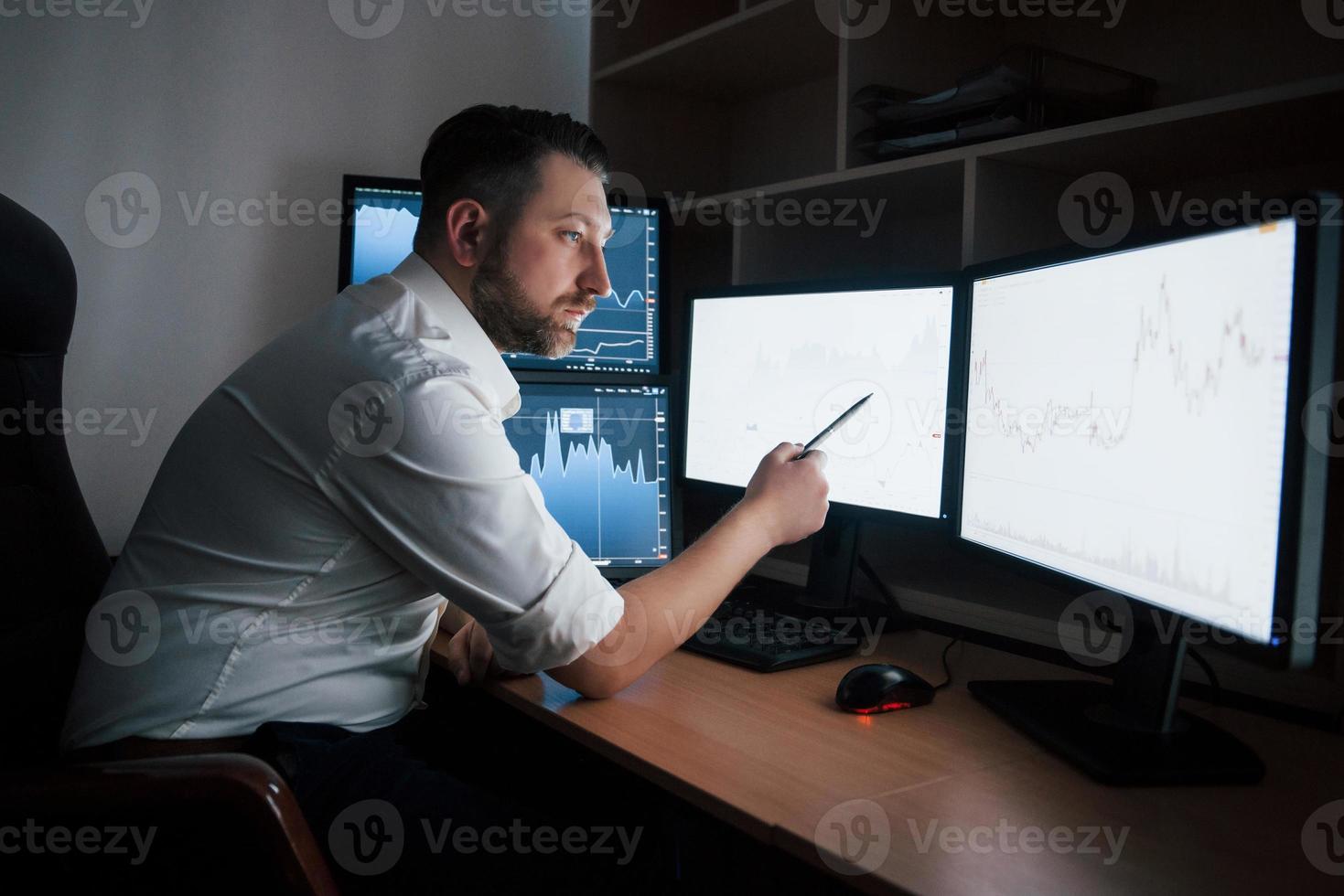 tantos monitores. hombre barbudo con camisa blanca trabaja en la oficina con múltiples pantallas de computadora en gráficos de índice foto