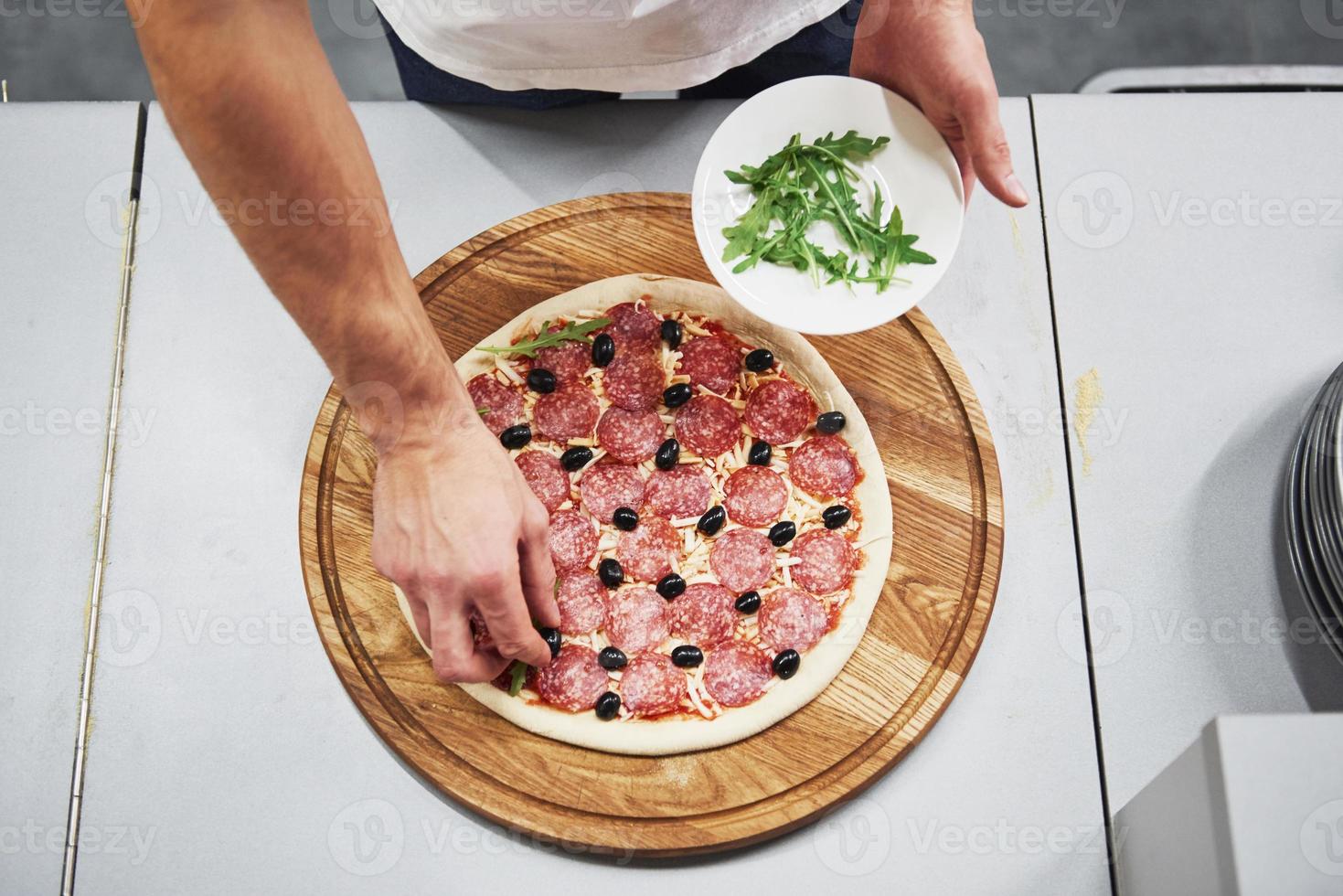 Work in progress. Top view of baker in white shirt putting olives to make delicious pizza for an order in restaurant photo