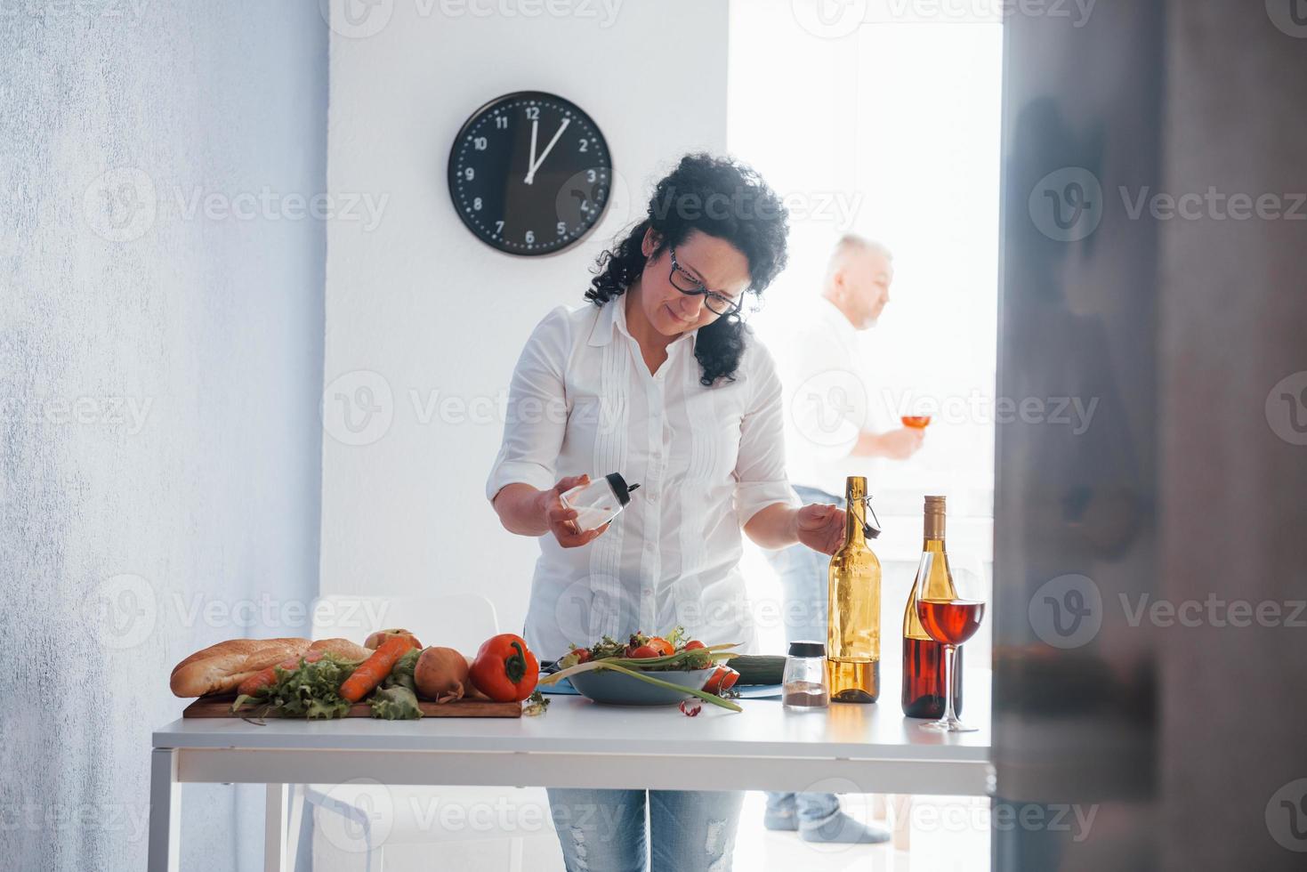 Spending time with wine near the window. Senior man and his wife in white shirt have romantic dinner on the kitchen photo