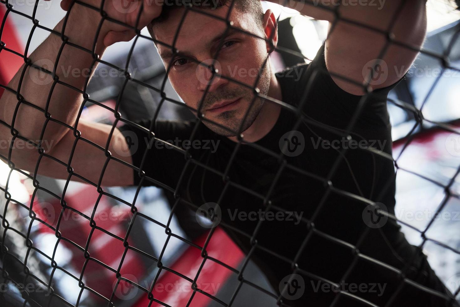 Strong man. Sportive guy in black shirt stands behind the fence in the room with daylight behind photo