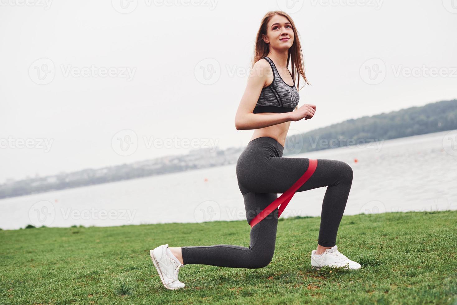 Side view. Fitness girl have morning workout at cloudy day near the lake with special red elastic band photo