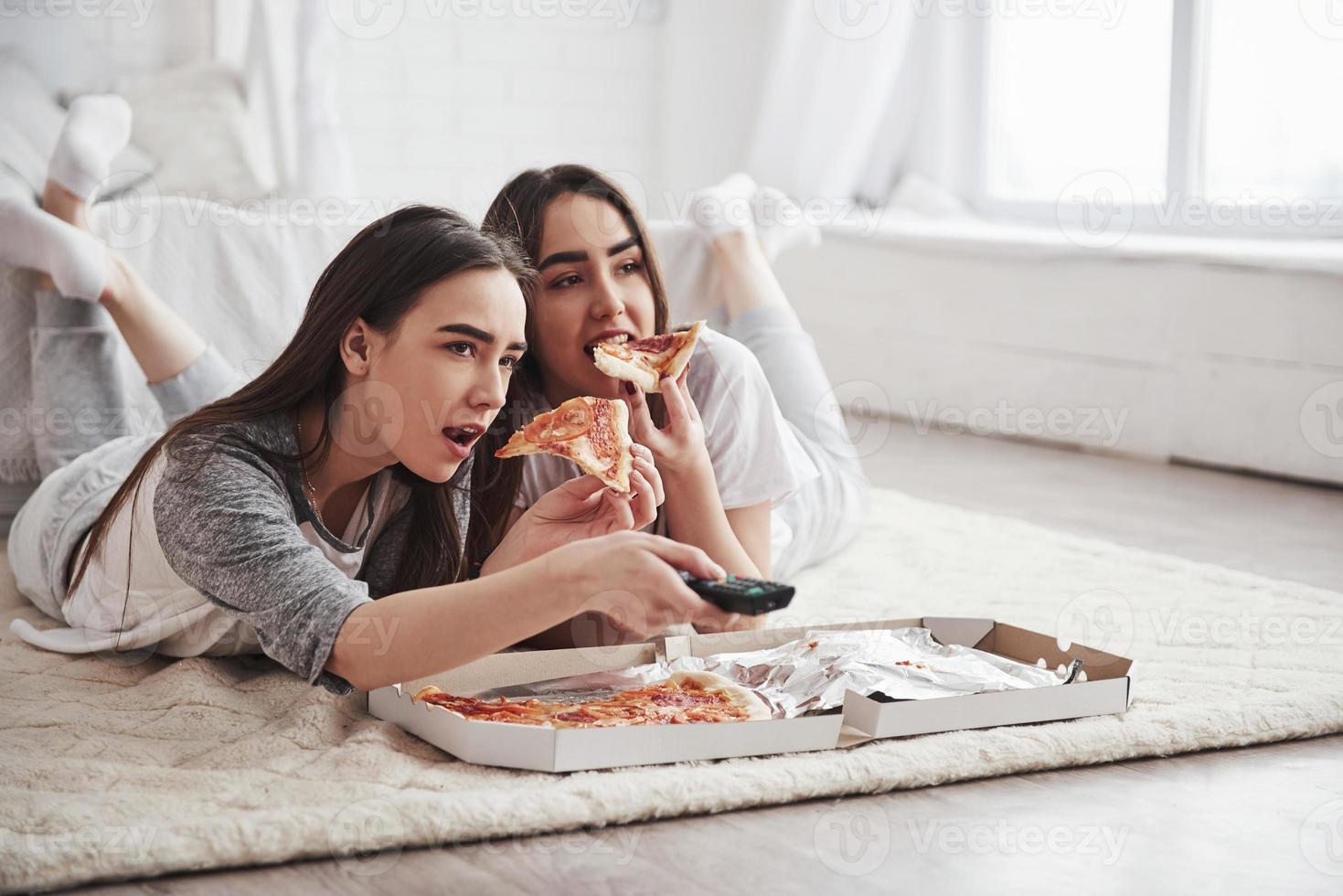Leisure time. Sisters eating pizza when watching TV while lying on the floor of beautiful bedroom at daytime photo