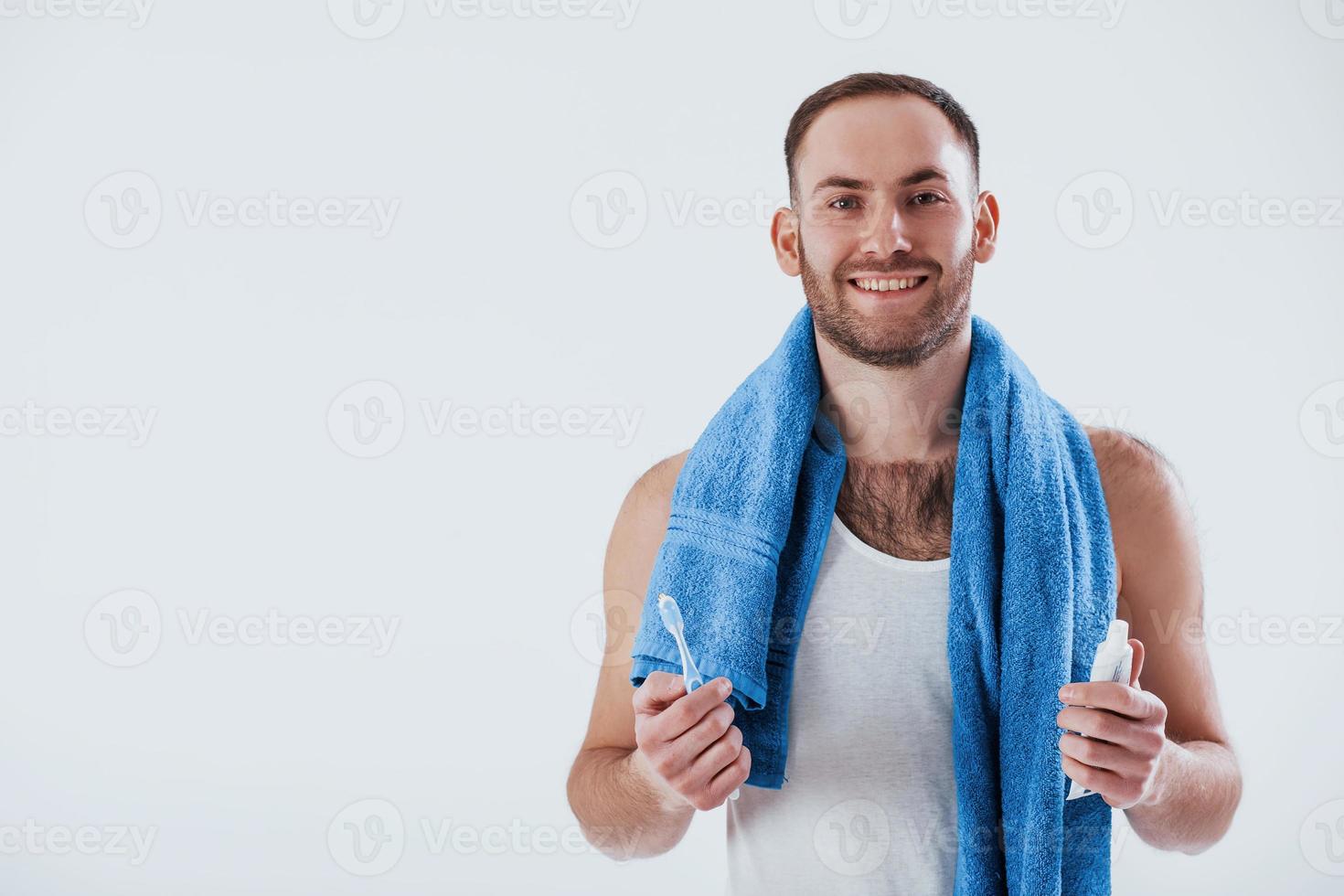 Toothbruh in hand. Man with blue towel stands against white background in the studio photo