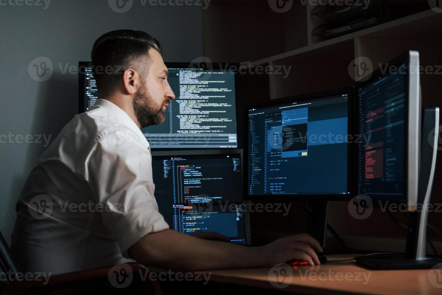 Adult experienced programmer. Bearded man in white shirt works in the office with multiple computer screens in index charts photo
