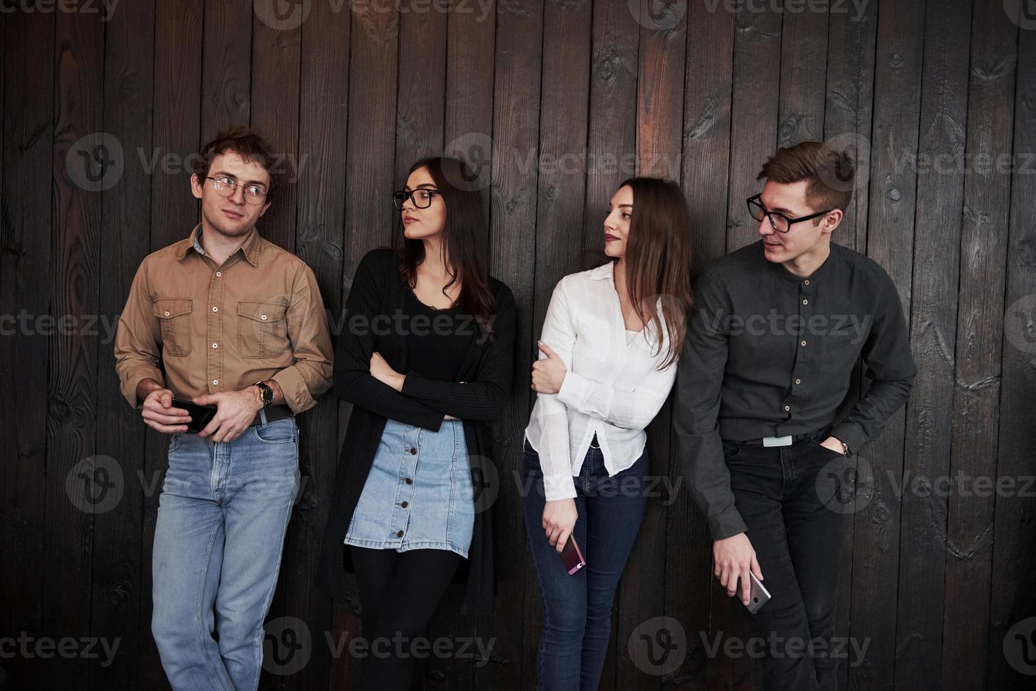 Looking at the guy on the left side. Youth stands against black wooden wall. Group of friends spending time together photo