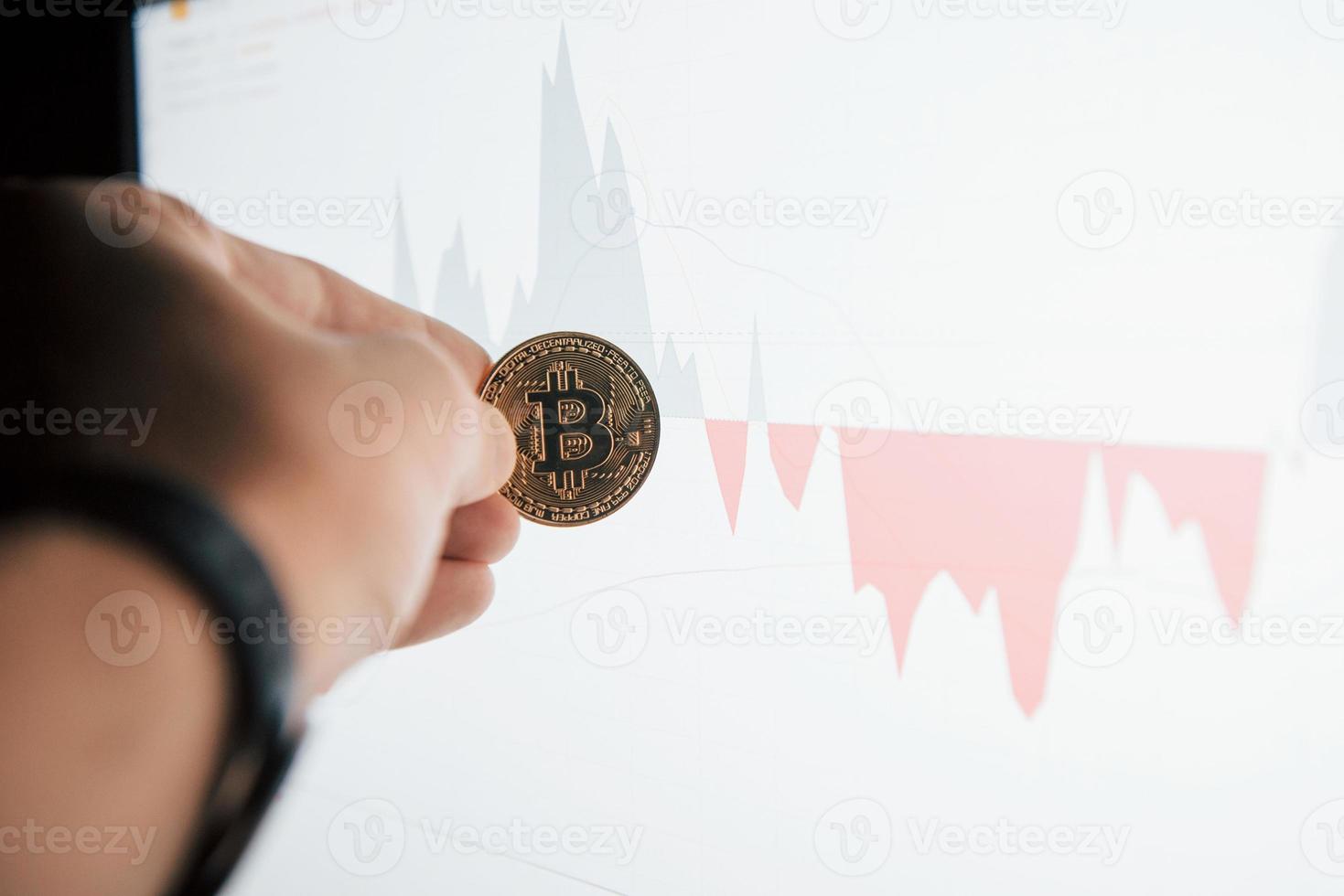 mano de hombre sosteniendo bitcoin en oficina moderna contra monitor con gráfico foto