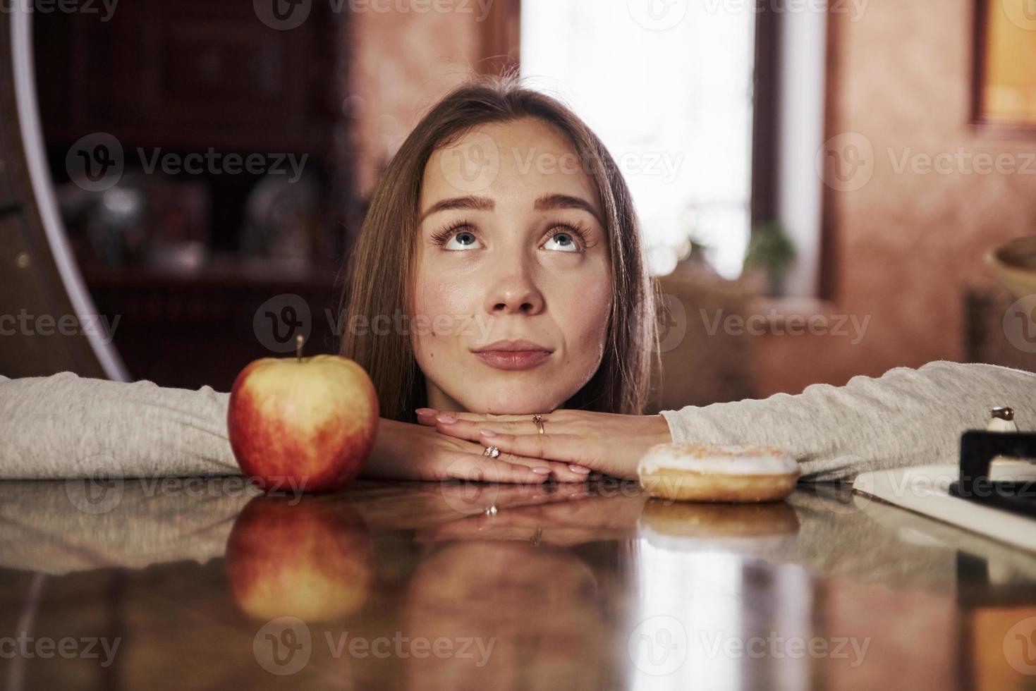 linda joven ama de casa apoyada en la mesa con manzana y galleta y mira hacia arriba foto