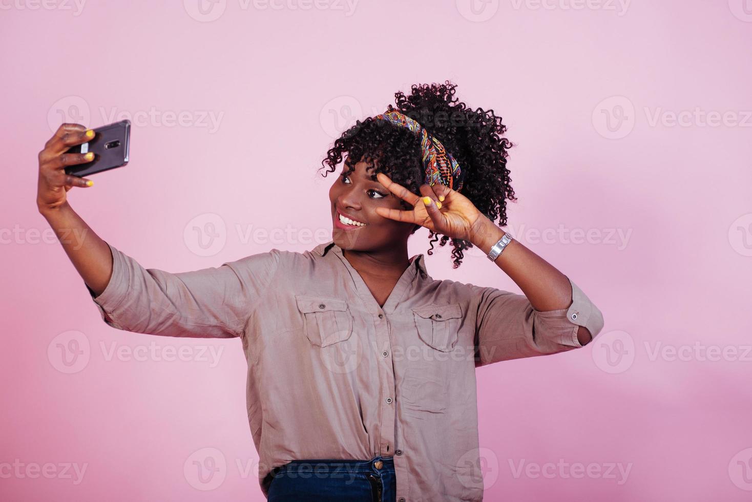 Photo of the day. Attractive afro american woman in casual clothes takes selfie and shows gesture with two fingers at pink background in the studio