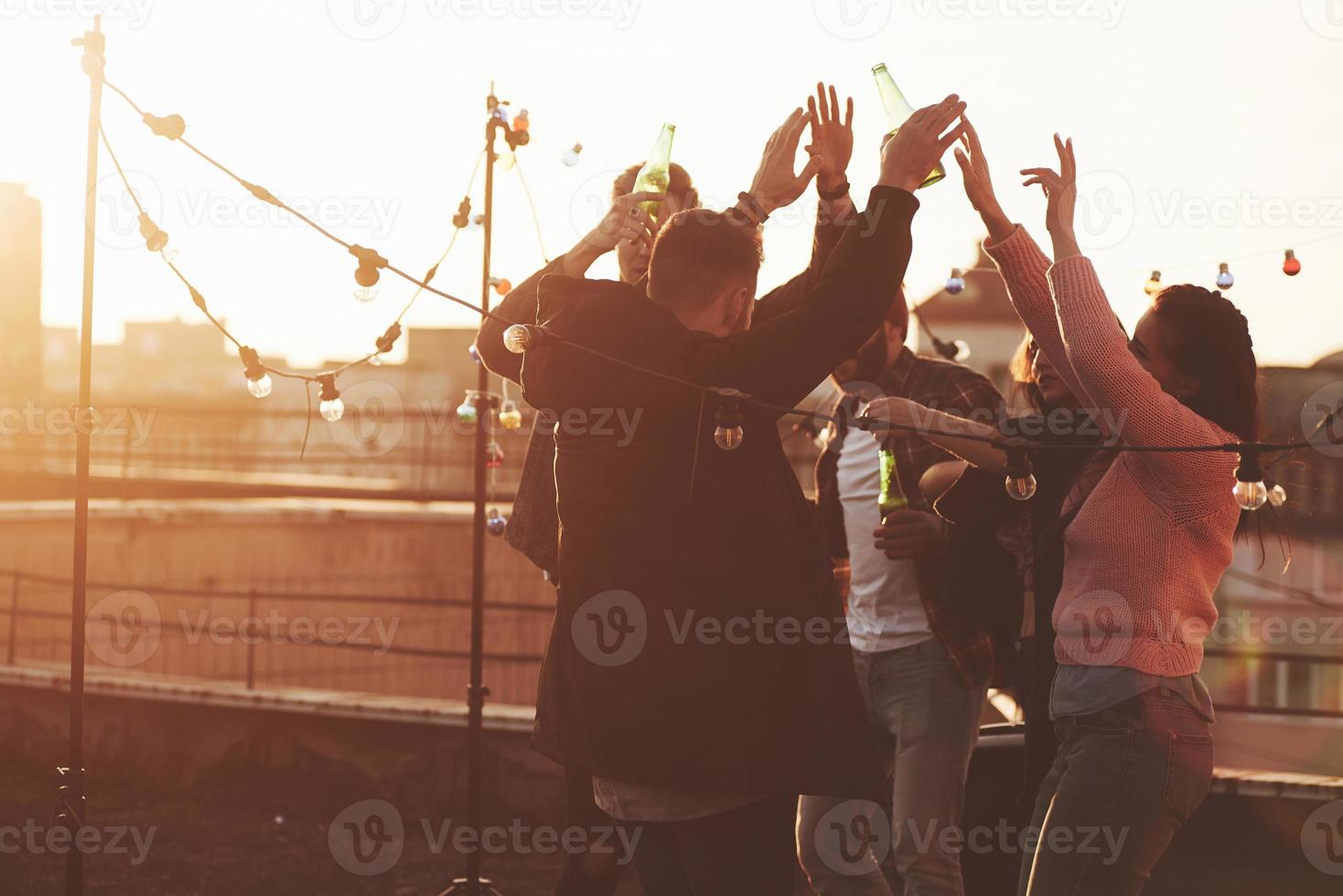 It's good weather. Holidays on the rooftop. Cheerful group of friends raised their hands up with alcohol photo