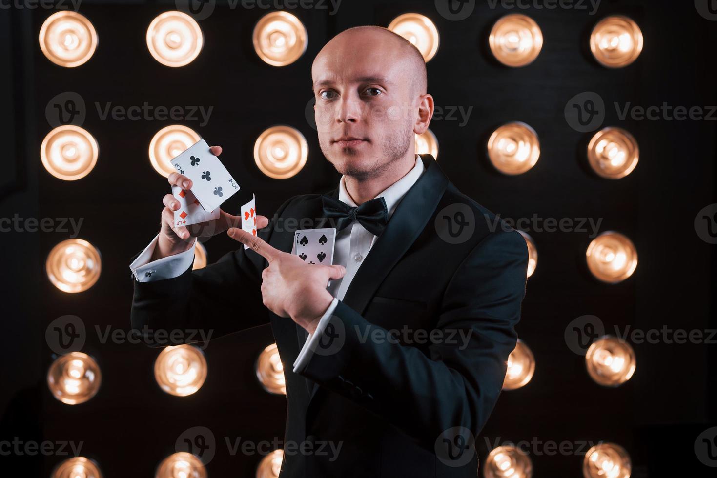 algunos trucos serios. mago en traje negro y con cartas de juego de pie en la habitación con iluminación especial en el backstage foto