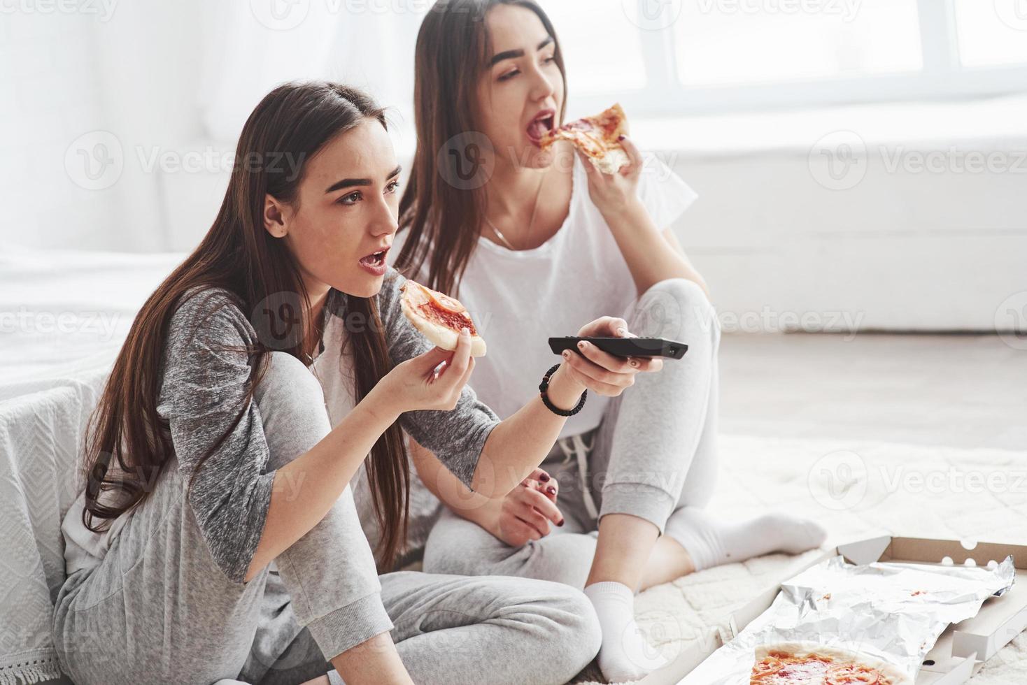 no puedo quitar los ojos de la película. hermanas comiendo pizza cuando ven la televisión mientras se sientan en el piso de un hermoso dormitorio durante el día foto